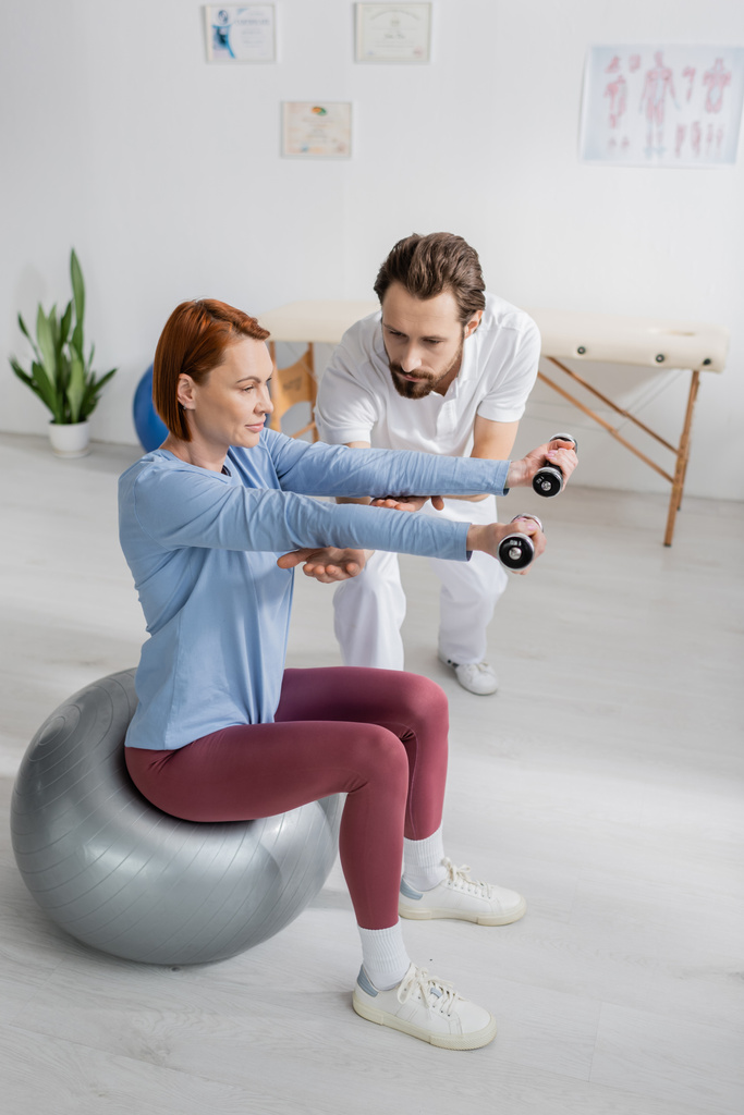 rehabilitológus segít a vörös hajú nőnek súlyzókkal edzeni, miközben fitnesz labdán ül a rehabilitációs központban. - Fotó, kép