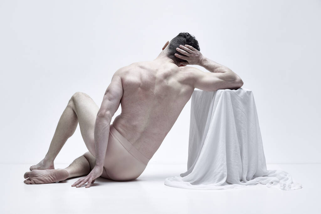 Achteraanzicht portret van gespierde shirtloze man zittend in ondergoed en poserend als standbeeld tegen witte studio achtergrond. Concept van mannelijke lichaamsesthetiek, mens schoonheid, inspiratie, kunst - Foto, afbeelding