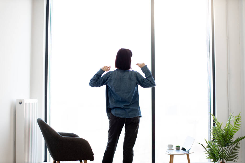 Вид на расслабленную брюнетку, растягивающую руки, стоя в просторном офисе рядом с панорамным окном. Молодой кавказский работник в повседневной одежде наслаждается кофе-брейком в рабочее время. - Фото, изображение