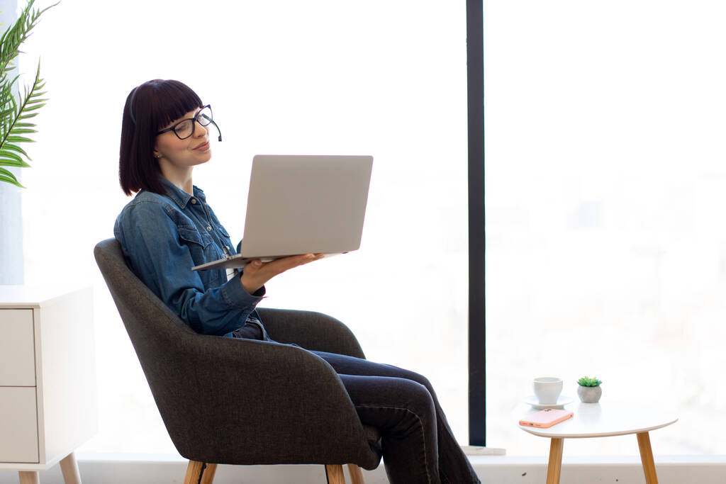 Elinde dizüstü bilgisayarla gözlüklü gülümseyen genç bir kadın akıllı telefonlu kahve masasının yanında oturuyor ve içiyor. Ofisteki kablosuz kulaklığı kullanarak çevrimiçi brifinge katılan etkili bir danışman.. - Fotoğraf, Görsel