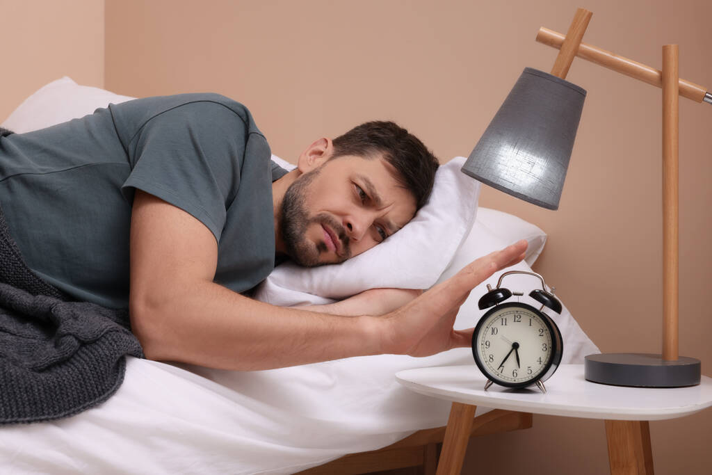 Сонний чоловік вимикає будильник на тумбочці вранці
 - Фото, зображення