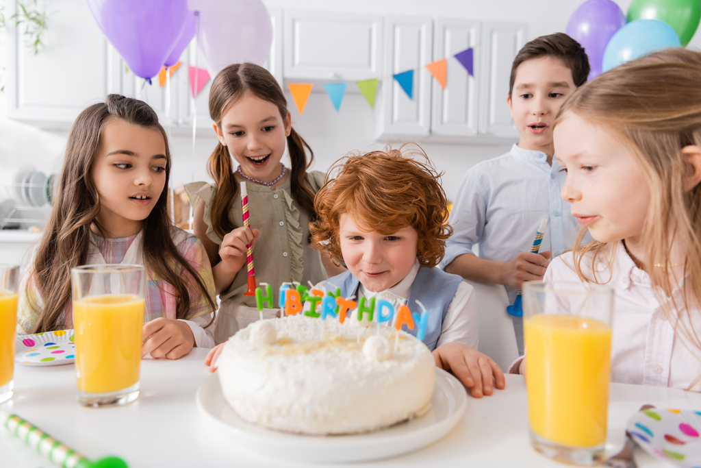 赤頭の男の子が家でパーティー中に友達の近くの誕生日ケーキにキャンドルを吹いて   - 写真・画像