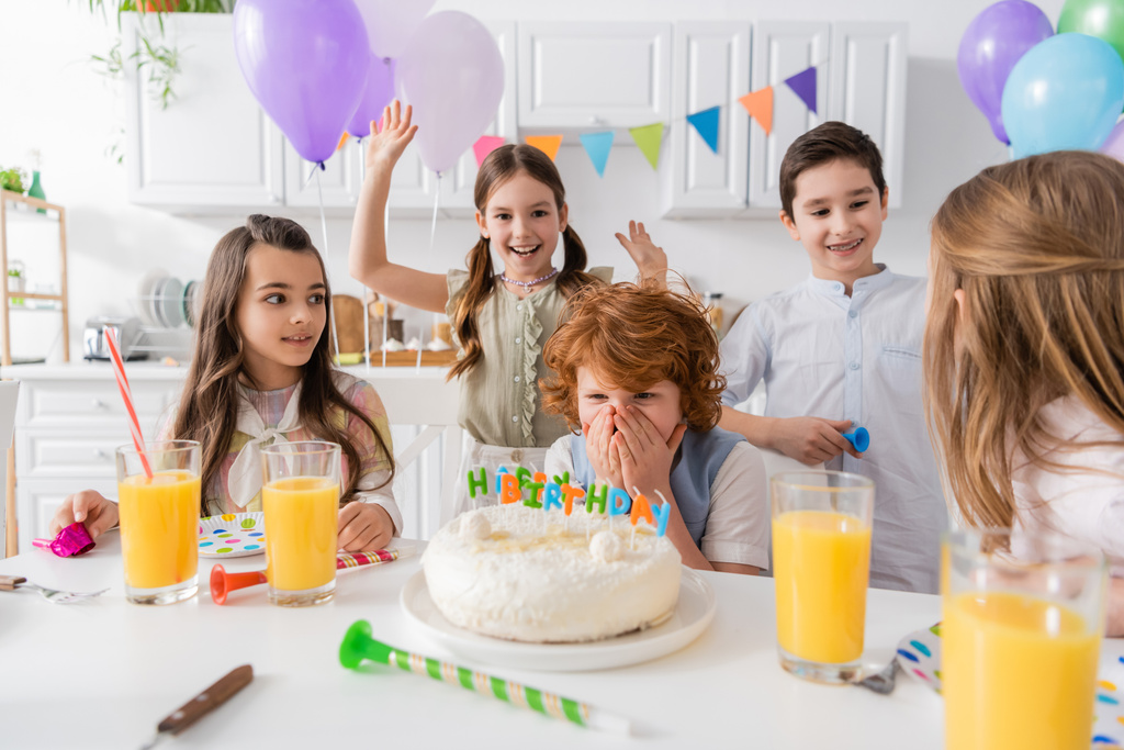 рудий хлопчик вкриває обличчя, дивлячись на торт на день народження поруч з друзями під час вечірки вдома
 - Фото, зображення