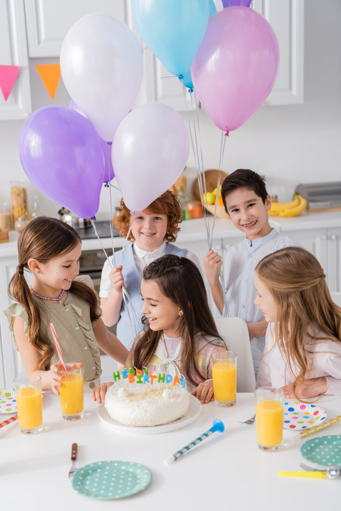 ケーキの近くの誕生日の女の子とテーブルの上のパーティーの角を見ている風船の明るい子供たち  - 写真・画像