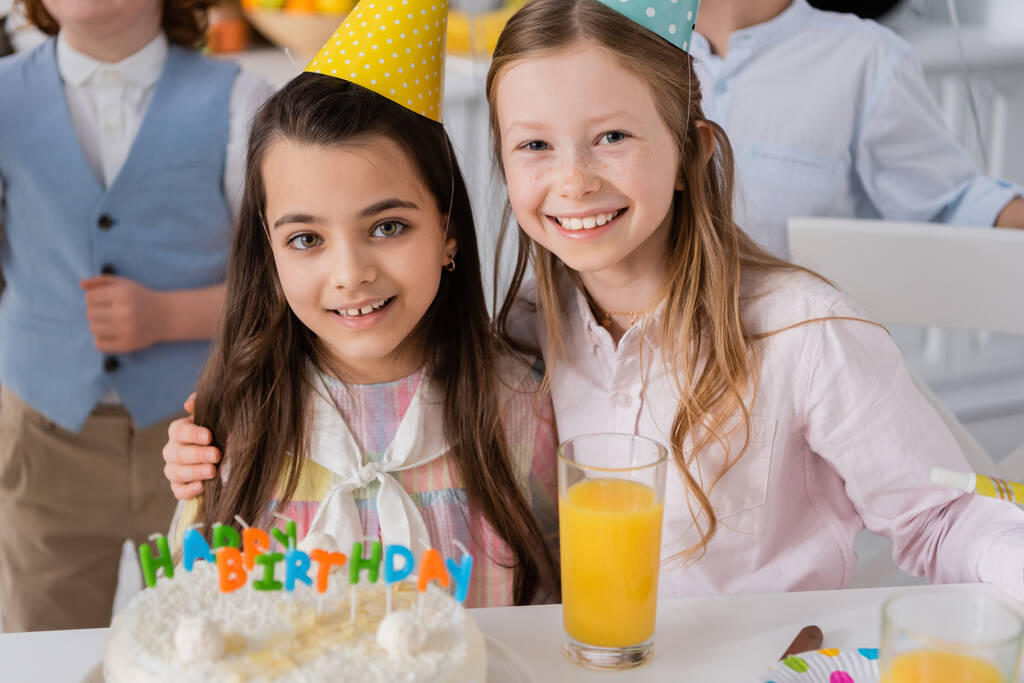счастливая девушка обнимает веселую подругу в шапочке для вечеринок рядом с праздничным тортом  - Фото, изображение