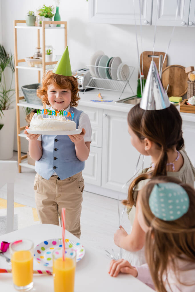 šťastný zrzka chlapec drží narozeninový dort se svíčkami v blízkosti přátel během párty doma   - Fotografie, Obrázek