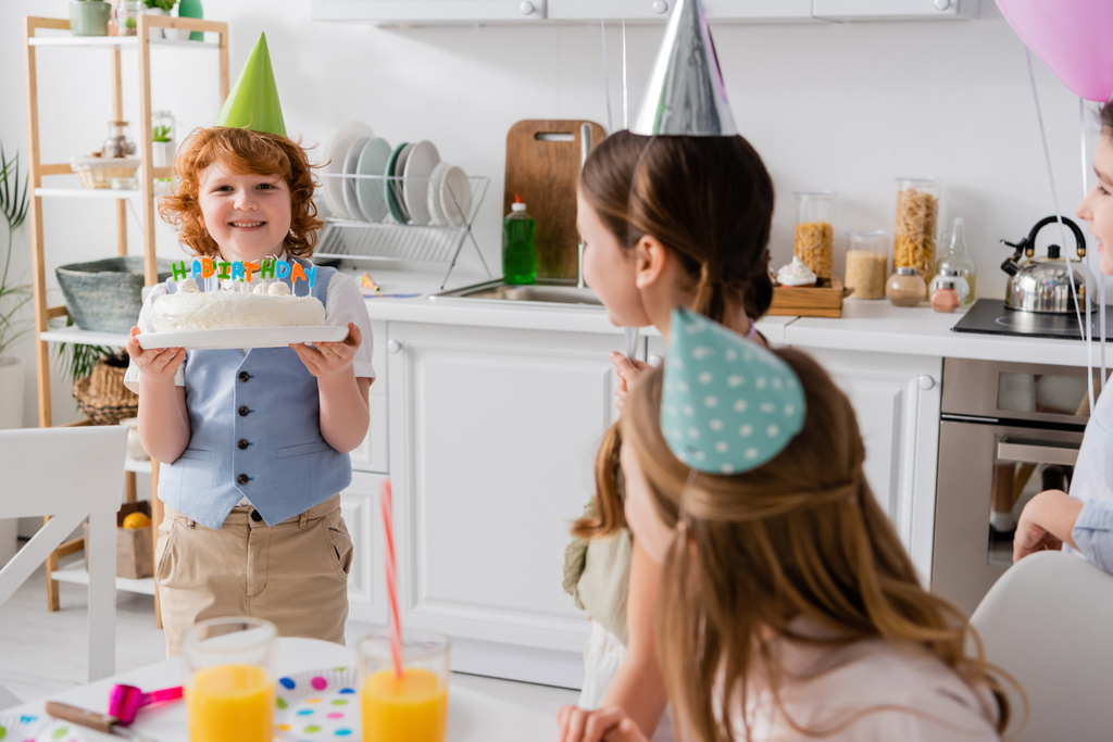 heureux rousse garçon tenant gâteau d'anniversaire avec des bougies près des amis joyeux pendant la fête à la maison  - Photo, image