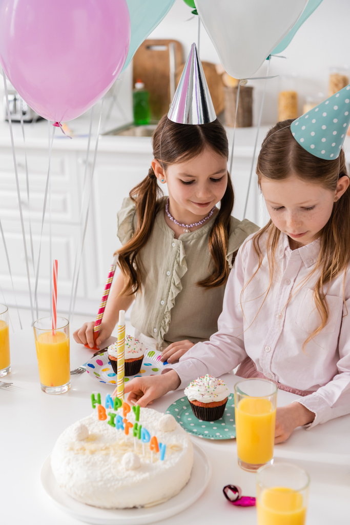 niñas preadolescentes en gorras de fiesta mirando pastel de cumpleaños y cupcakes en la mesa  - Foto, imagen
