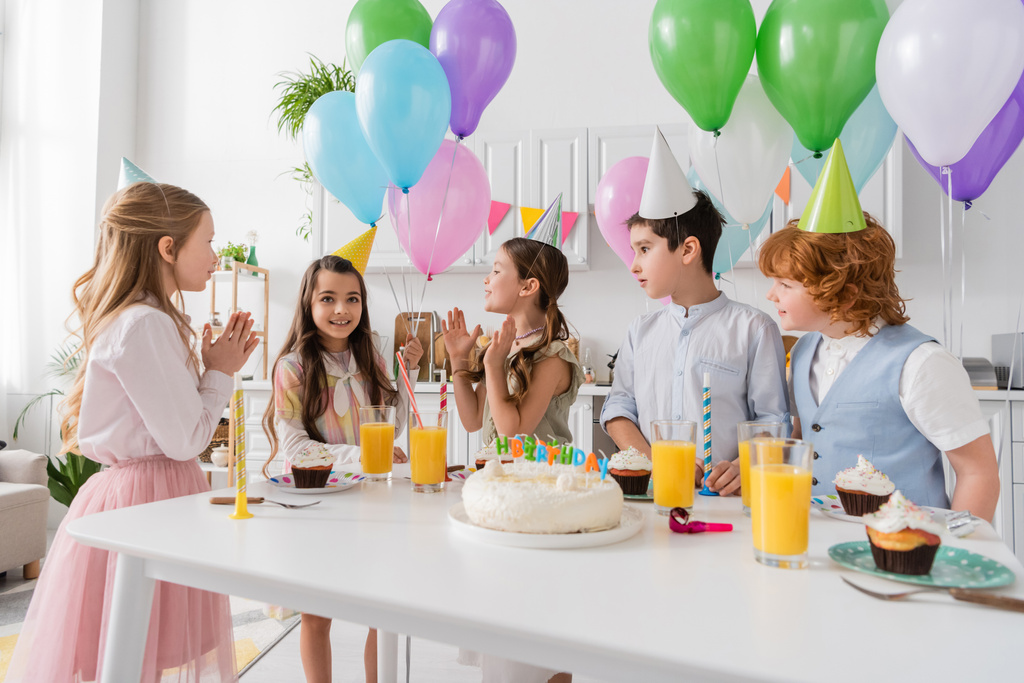 счастливые дети хлопают в ладоши и поют счастливую песню дня рождения рядом с тортом со свечами и воздушными шарами  - Фото, изображение