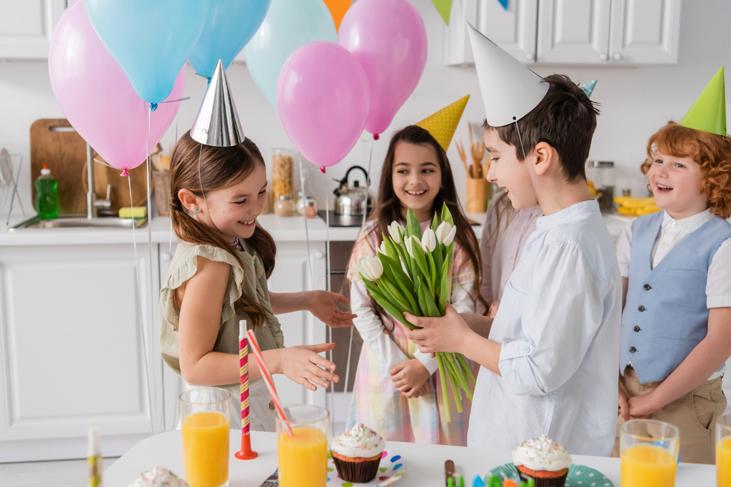 мальчик-подросток дарит тюльпаны веселой имениннице рядом с друзьями на размытом фоне  - Фото, изображение