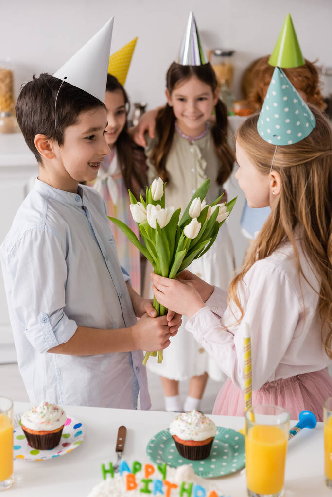 preteen boy with braces giving tulips to cheerful birthday girl near friends on blurred background  - Photo, Image