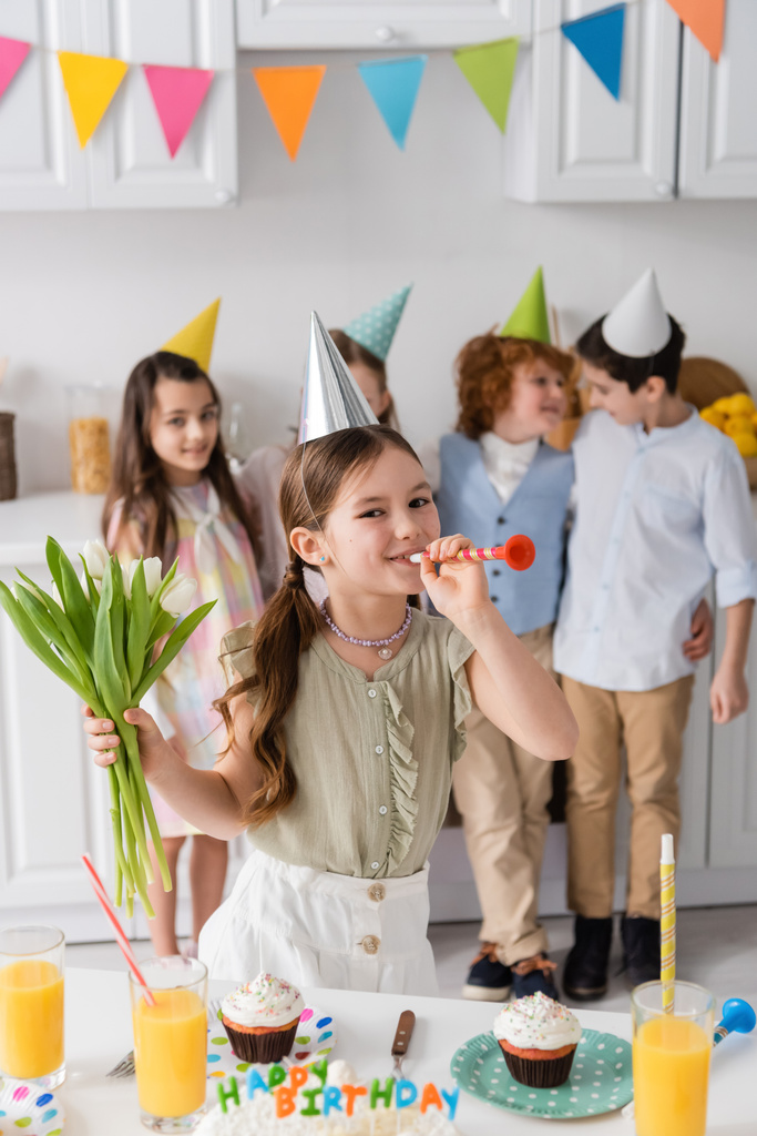 menina alegre segurando tulipas e soprando chifre de festa perto de amigos durante a festa de aniversário em casa  - Foto, Imagem