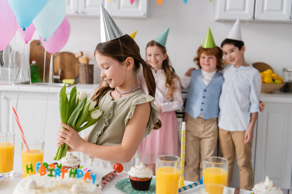 veselá dívka ve straně čepice drží tulipány vedle přátel během narozeninové párty doma  - Fotografie, Obrázek