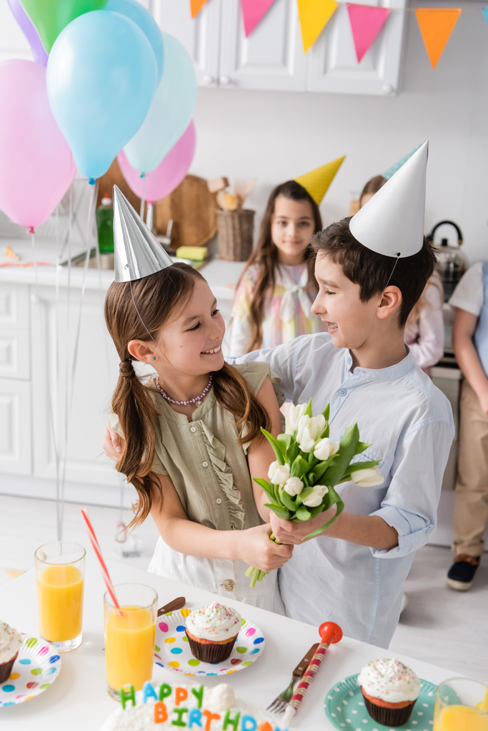 szczęśliwa dziewczyna w party cap gospodarstwa tulipany w pobliżu chłopca i obok przyjaciół podczas urodzin w domu  - Zdjęcie, obraz