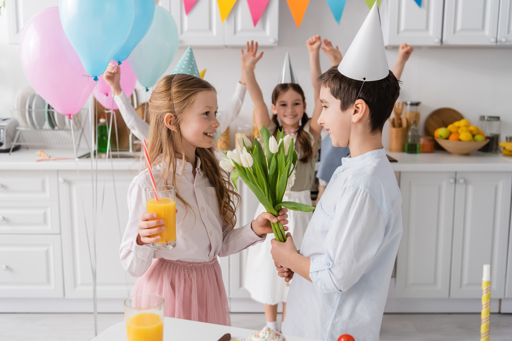 gelukkig meisje in party cap het nemen van tulpen van jongen in de buurt vrienden op wazig achtergrond  - Foto, afbeelding