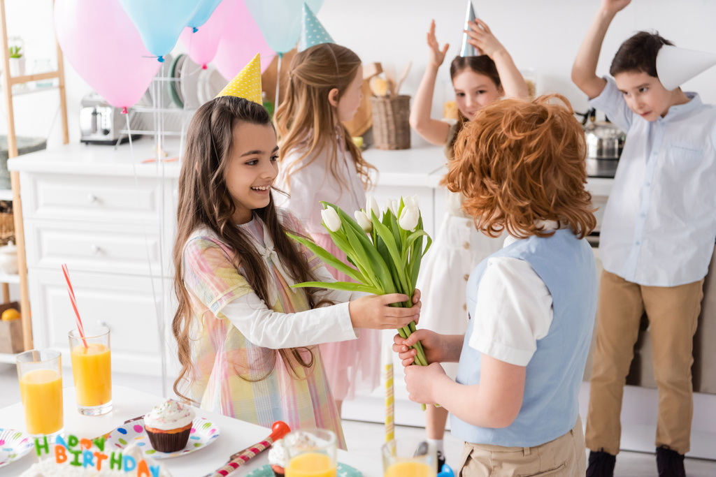 menina feliz em boné de festa levando tulipas de menino ruivo perto de amigos no fundo borrado  - Foto, Imagem