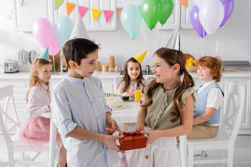 happy preteen boy giving present to cheerful birthday girl near friends on blurred background  - Photo, Image