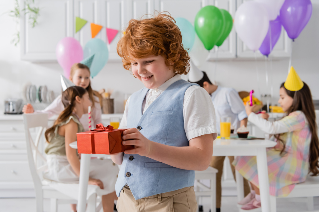 vidám vörös hajú fiú tartja születésnapi ajándék közel barátok alatt party otthon  - Fotó, kép