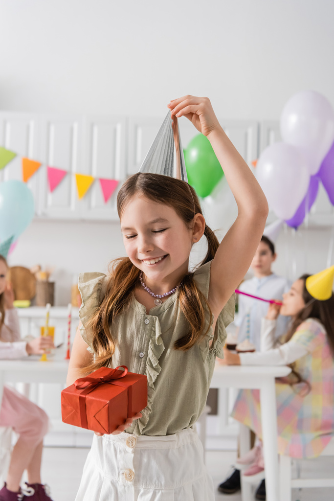 веселая девушка регулирует крышку вечеринки и держит подарок на день рождения рядом с друзьями на размытом фоне  - Фото, изображение