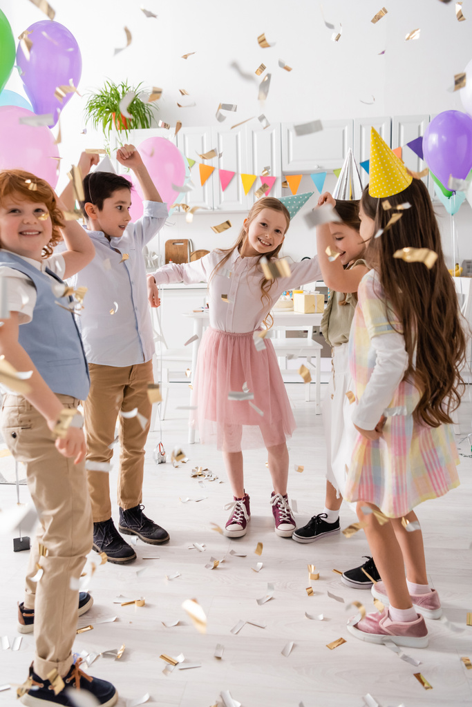 счастливые дети в шапочках для вечеринок танцуют под падающими конфетти во время празднования дня рождения дома  - Фото, изображение