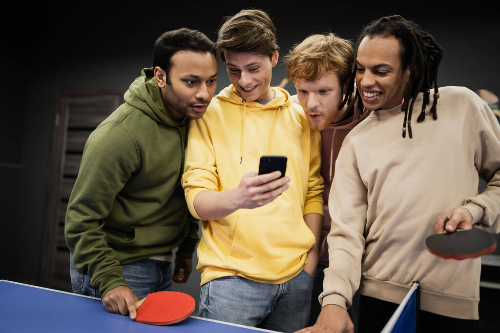 Vrolijke multi-etnische vrienden met behulp van mobiele telefoon in de buurt van tafeltennis in gaming club  - Foto, afbeelding