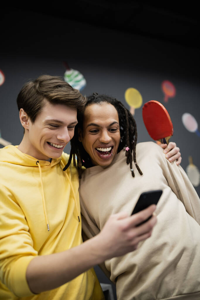 Glimlachende man met behulp van smartphone en het houden van tennis racket in de buurt van multiraciale vriend in gaming club  - Foto, afbeelding