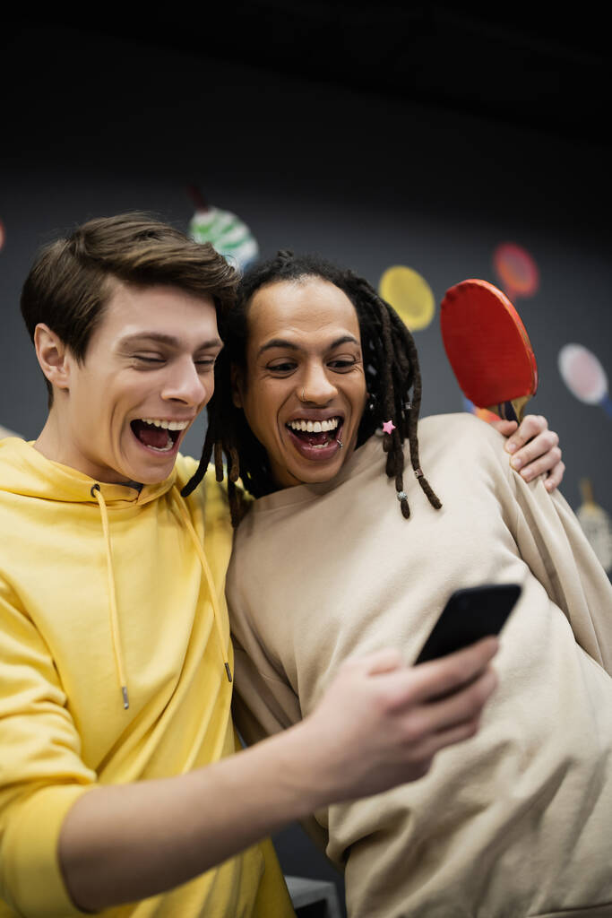 Positieve interraciale vrienden met tennis racket met behulp van mobiele telefoon in gaming club  - Foto, afbeelding