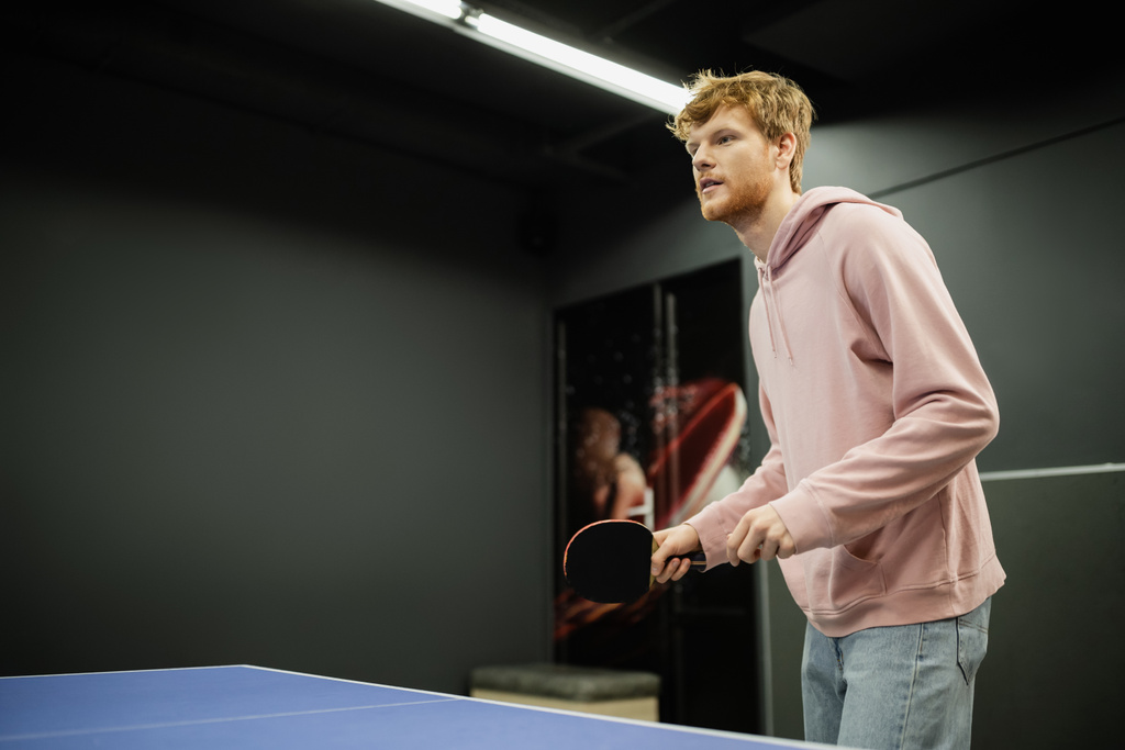 Jovem ruiva homem segurando raquete enquanto joga tênis de mesa no clube de jogos  - Foto, Imagem