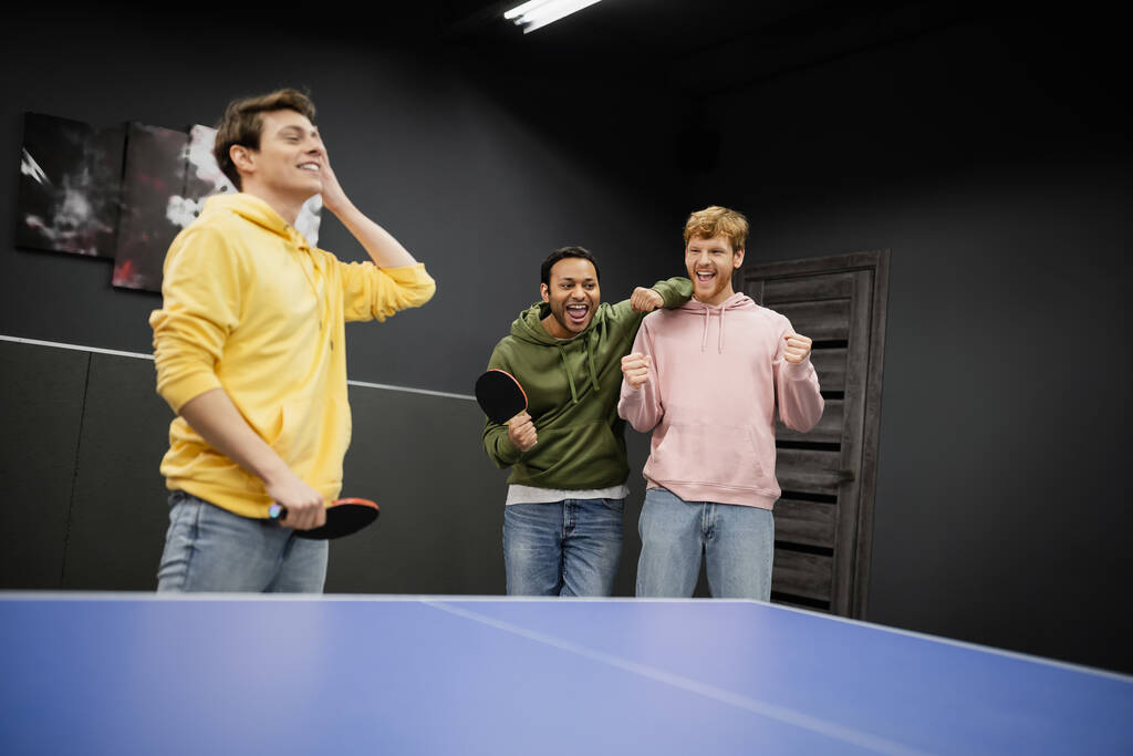 Podekscytowani międzyrasowi mężczyźni stojący w pobliżu niewyraźnego przyjaciela z rakietą tenisową w klubie gier  - Zdjęcie, obraz