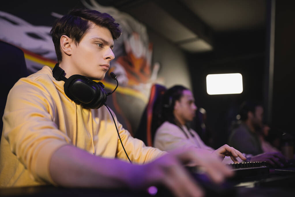 Mladý muž se sluchátky hrát videohry na počítači v blízkosti rozmazaných přátel v kybernetickém klubu  - Fotografie, Obrázek