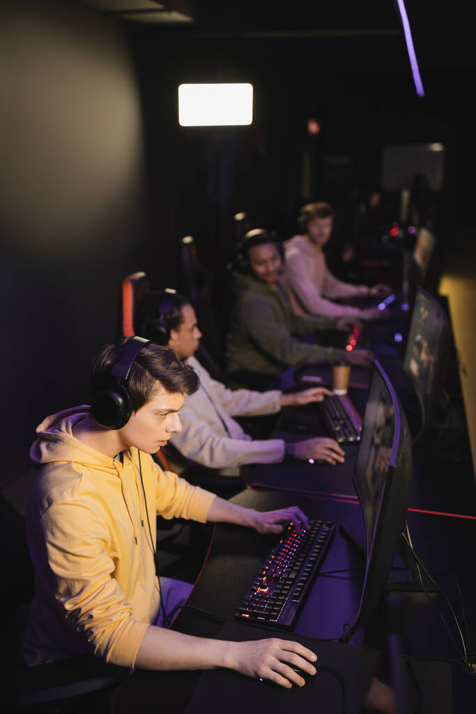 Spieler mit Kopfhörer spielt Videospiel auf Computer in der Nähe verschwommenes gemischtes Team in Gaming-Club  - Foto, Bild