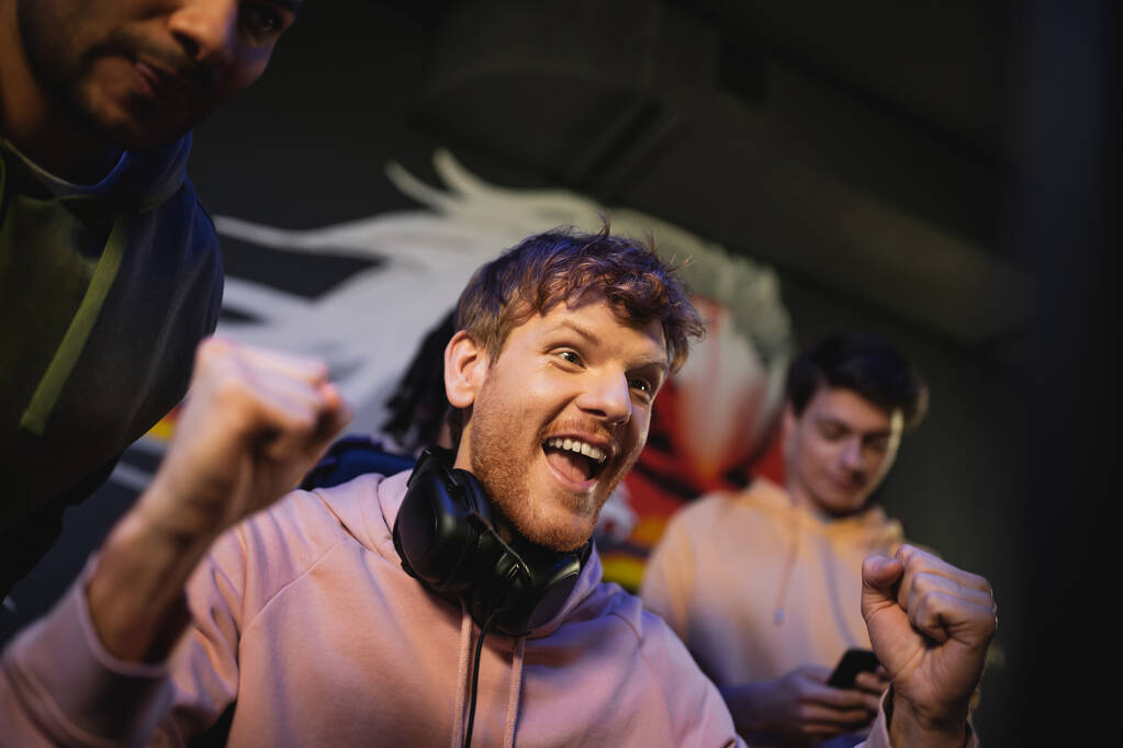 Excited man with headphones standing near interracial friends in gaming club  - Photo, Image