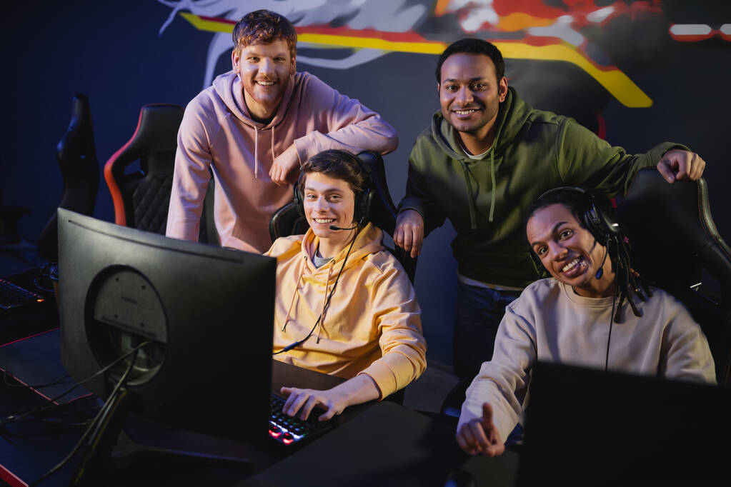Позитивные мультикультурные друзья в наушниках смотрят в камеру рядом с компьютерами в игровом клубе  - Фото, изображение