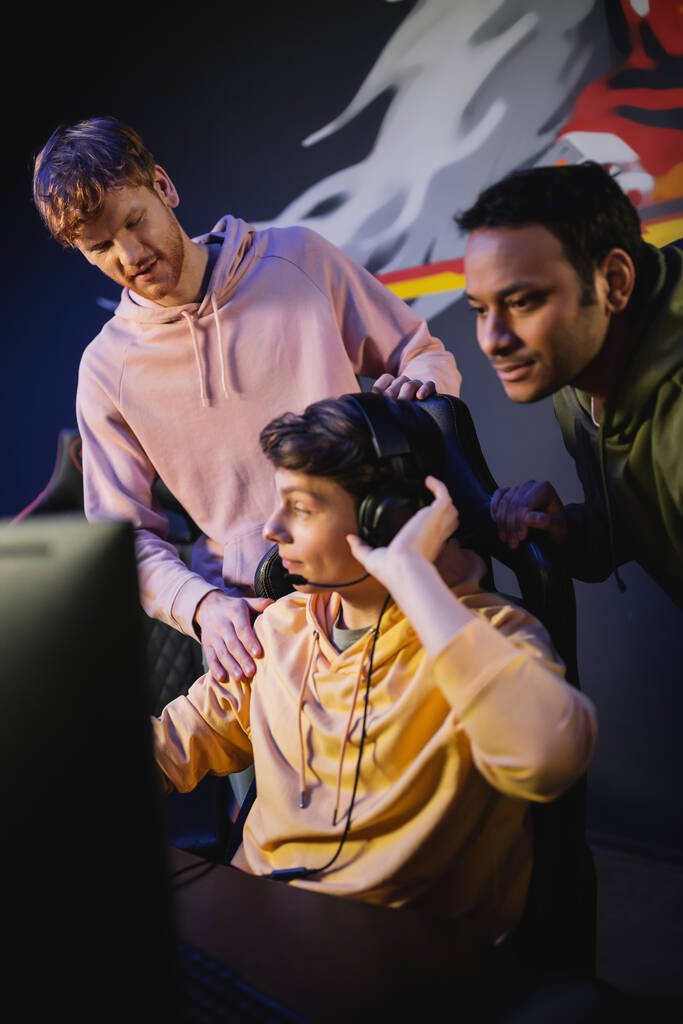 Jongeman met koptelefoon in de buurt van interraciale vrienden en computer in gaming club  - Foto, afbeelding
