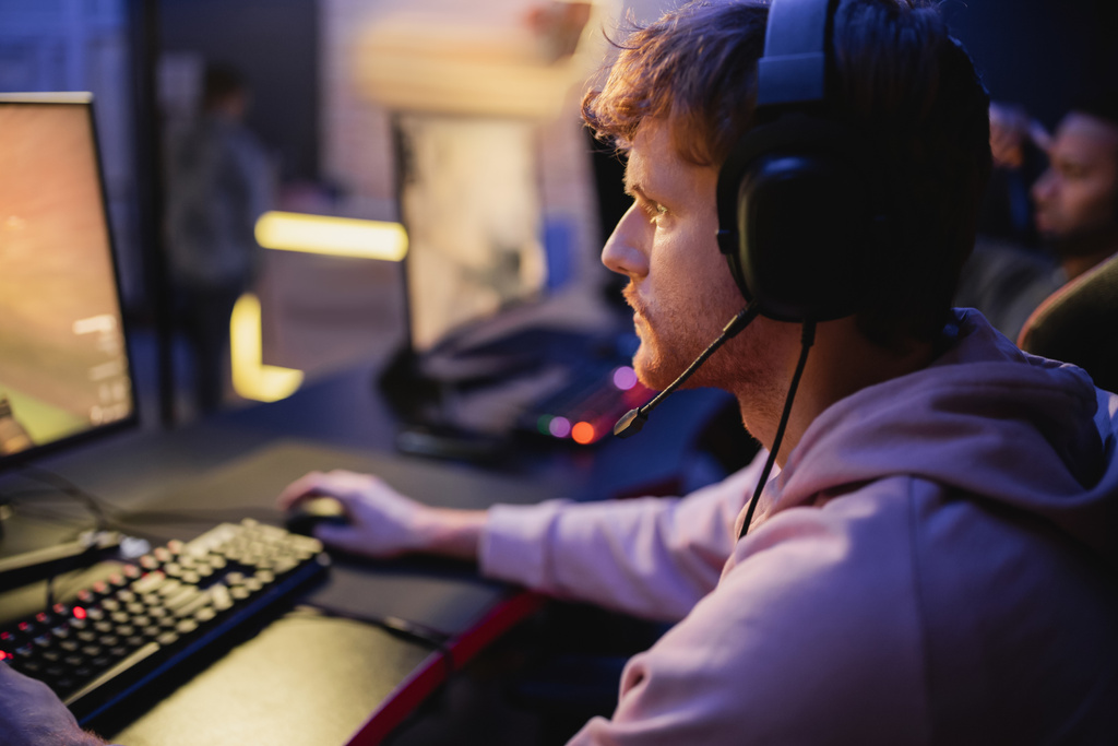 Seitenansicht des fokussierten Spielers in Kopfhörern, der ein Videospiel auf dem Computer in einem Spielclub spielt  - Foto, Bild