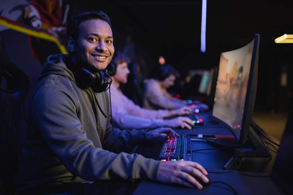 Lächelnder indischer Gamer blickt in Cyber-Club in Computernähe auf Kamera  - Foto, Bild