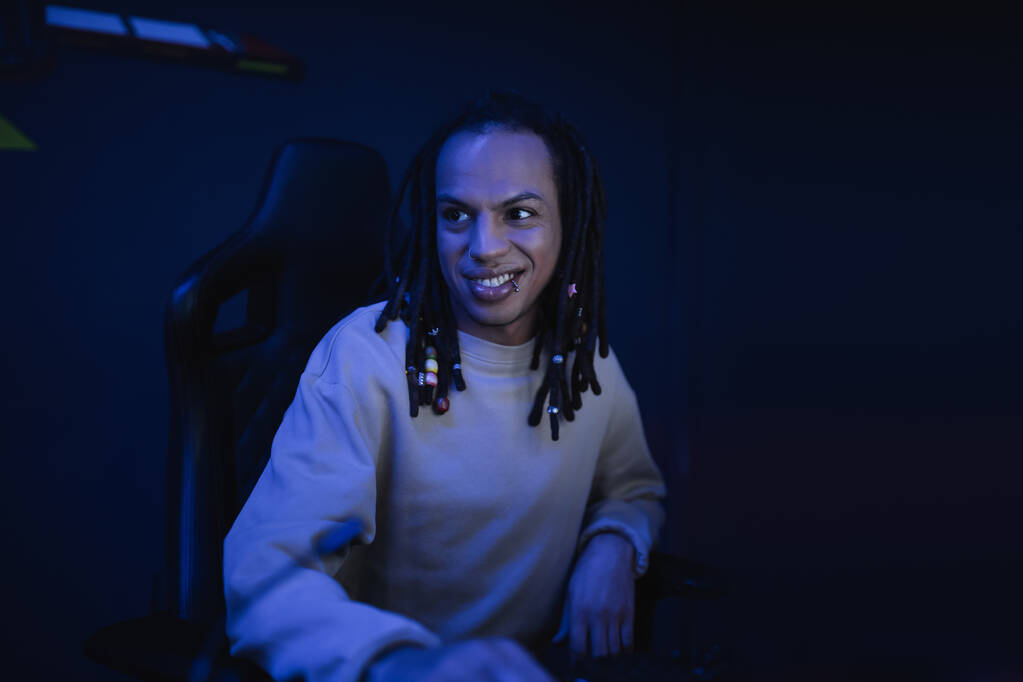 Jugador multirracial sonriente con rastas sentado en el club cibernético con iluminación  - Foto, Imagen