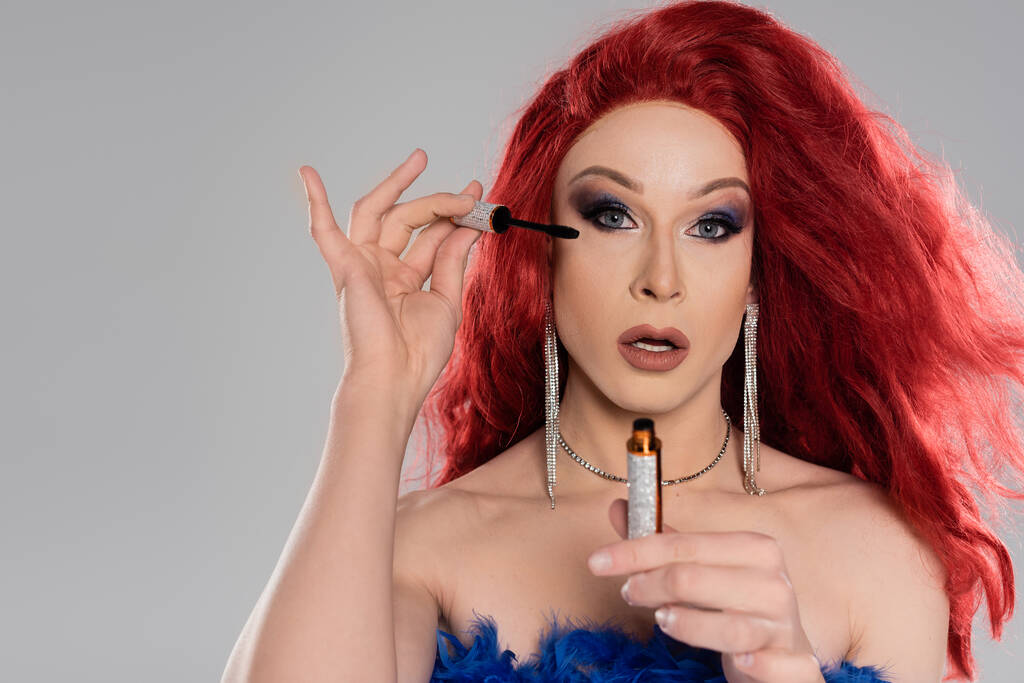Πορτρέτο της κομψής drag queen σε κόκκινη περούκα κρατώντας μάσκαρα και κοιτάζοντας κάμερα που απομονώνονται σε γκρι - Φωτογραφία, εικόνα