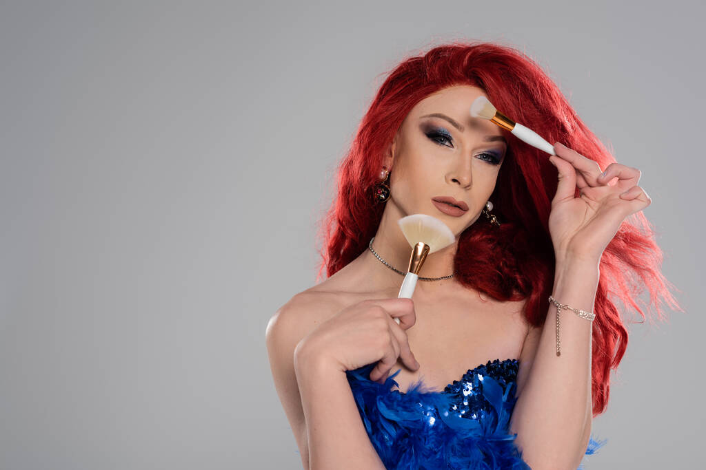 Портрет элегантного трансвестита, держащего косметические кисти возле лица, изолированного на сером   - Фото, изображение
