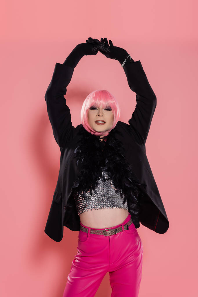 Lächelnde Drag Queen in Jacke mit Federn und Make-up posiert auf rosa Hintergrund  - Foto, Bild