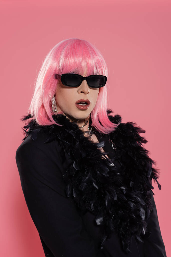 Retrato de persona transgénero con estilo con maquillaje y gafas de sol posando en chaqueta con plumas sobre fondo rosa  - Foto, Imagen
