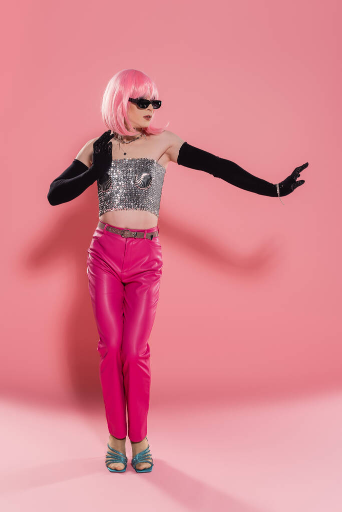 Pełna długość drag queen w błyszczącej bluzce i rękawiczkach pozujących na różowym tle  - Zdjęcie, obraz