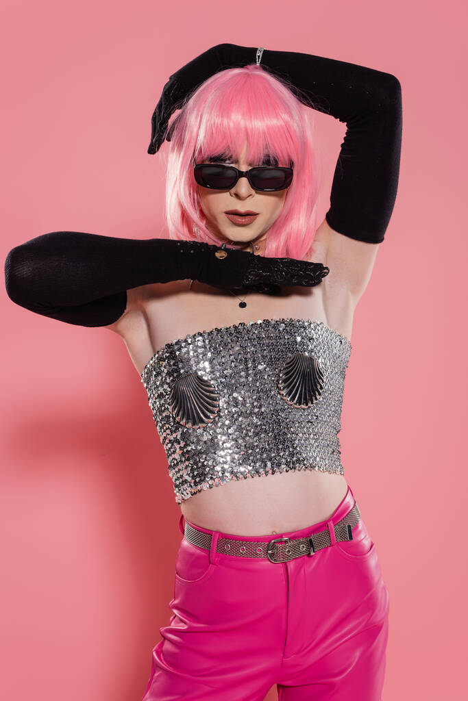 Retrato de drag queen en gafas de sol y top plateado brillante posando sobre fondo rosa  - Foto, imagen