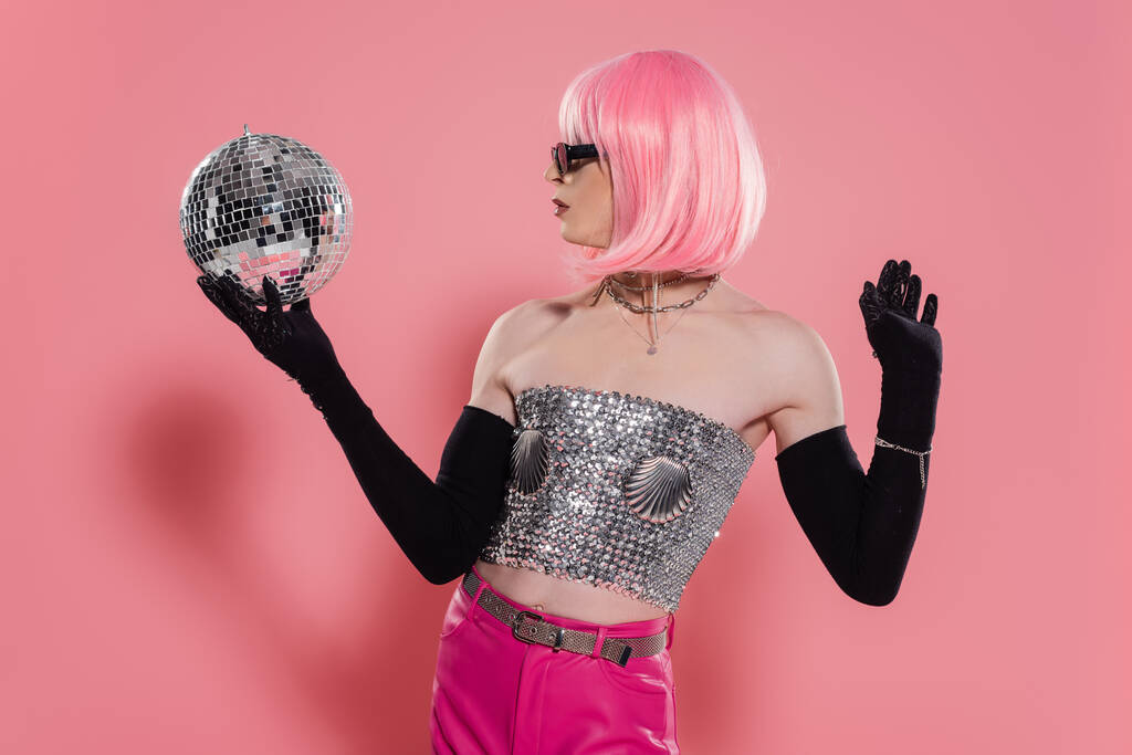 Boční pohled na trendy drag queen v jiskřivé horní a rukavice držící disco míč na růžovém pozadí  - Fotografie, Obrázek