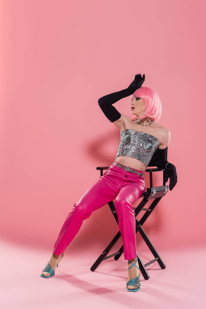ファッショナブルなドラッグクイーンのフル長さで光沢のあるトップポージングオン椅子上のピンクの背景  - 写真・画像