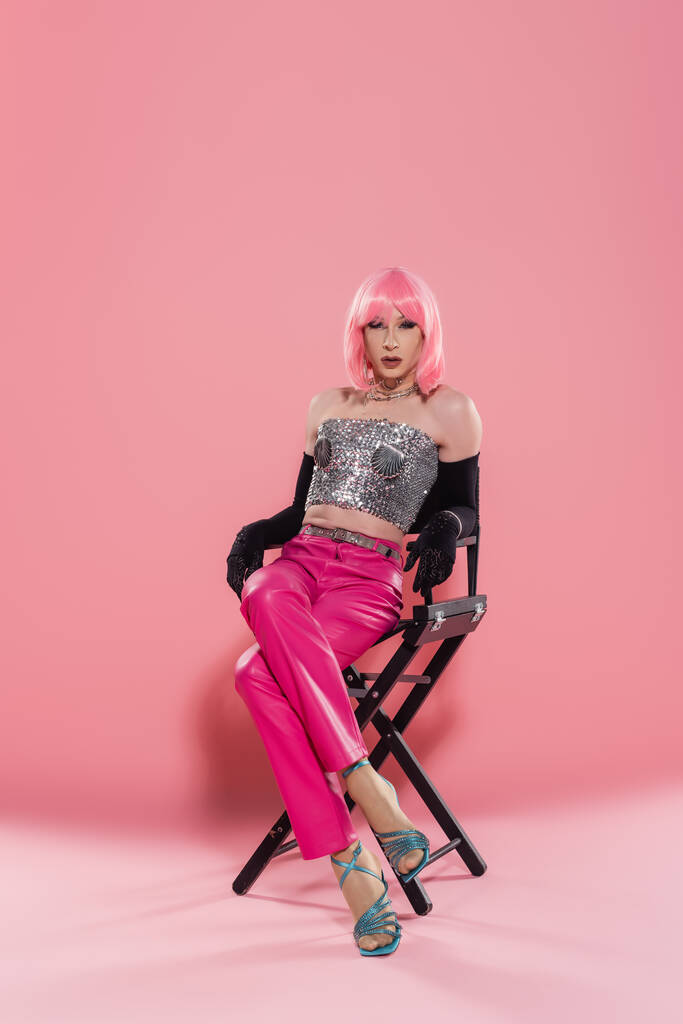 Fashionable drag queen in top and gloves posing while sitting on chair on pink background  - Photo, Image