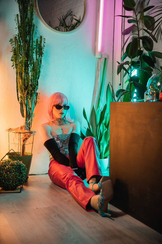 Stílusos transznemű személy napszemüvegben ül a padlón növények közelében otthon  - Fotó, kép