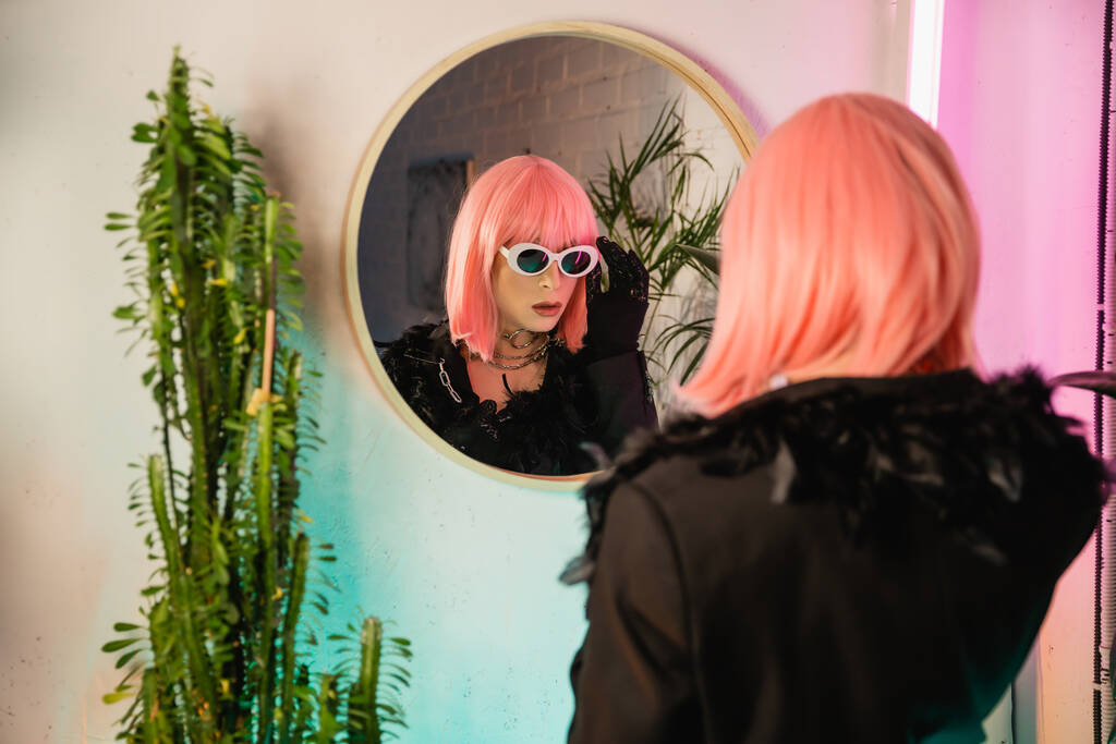 Trendy drag queen σε περούκα και γυαλιά ηλίου στέκεται κοντά στον καθρέφτη και τα φυτά στο σπίτι  - Φωτογραφία, εικόνα