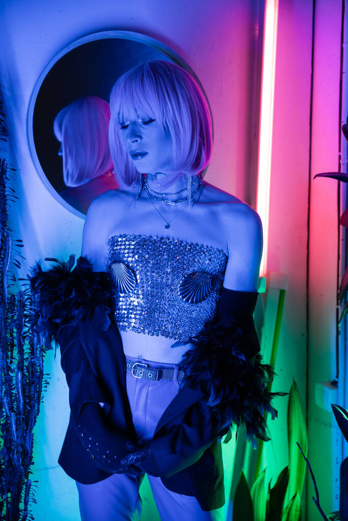 Drag queen en haut pétillant et veste debout dans la lumière au néon à la maison  - Photo, image