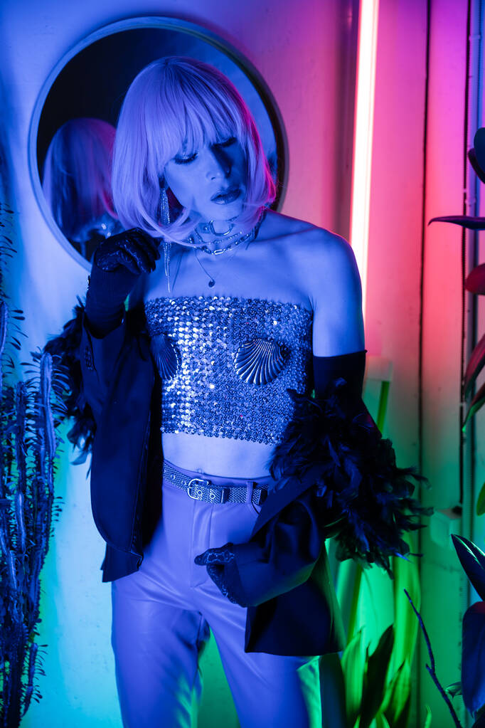 Modna transseksualistka w peruce i kurtce stojąca w pobliżu neonowego światła w domu  - Zdjęcie, obraz